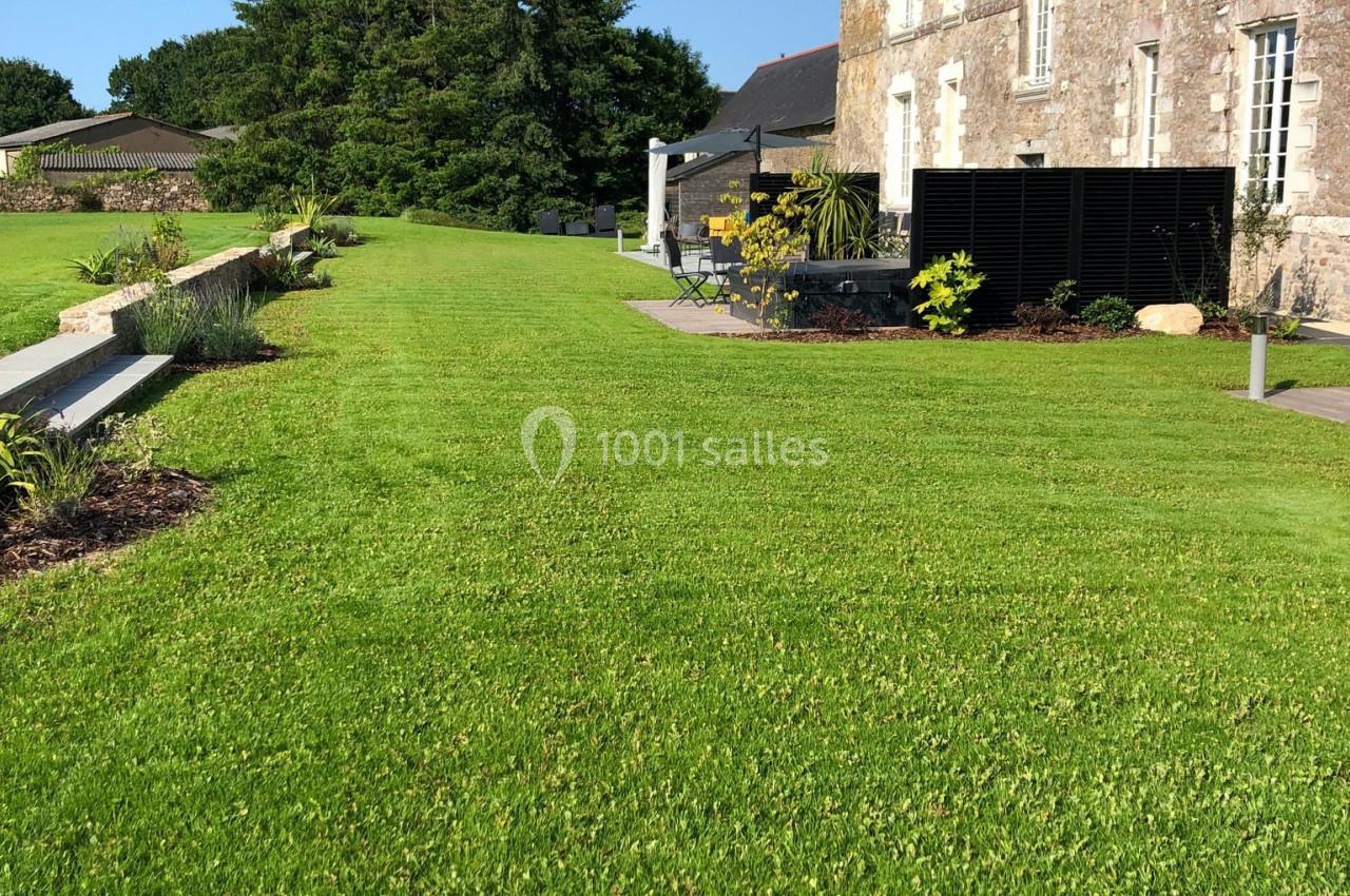 Pelouse bien entretenue devant une maison en pierre avec terrasse et arbres en arrière-plan.