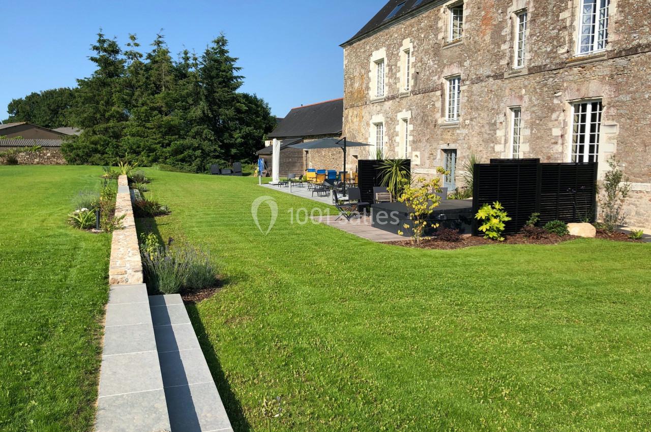 Jardin avec pelouse bien entretenue, terrasse aménagée et façade en pierre d'une maison ancienne sous un ciel dégagé.