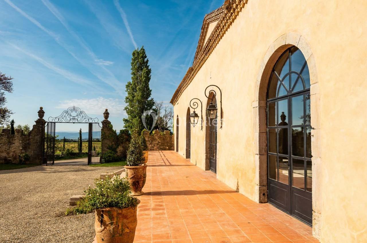 Façade en pierre d'un bâtiment avec grandes fenêtres cintrées, terrasse en terre cuite et vue sur un portail ouvragé.