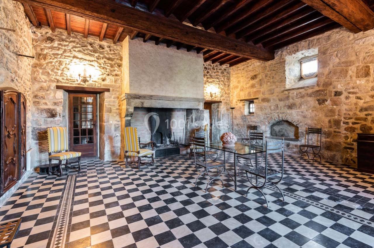 Salle rustique avec murs en pierre, cheminée, plafond en bois, sol à damier noir et blanc, et mobilier en métal.