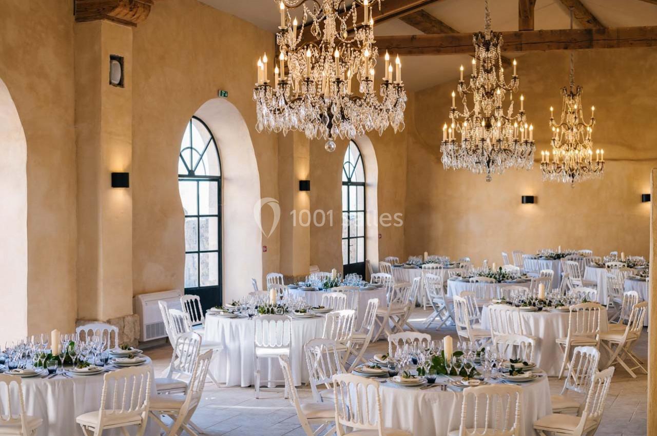 Salle de réception élégante avec murs beige, grandes fenêtres cintrées, lustres en cristal et tables rondes dressées.
