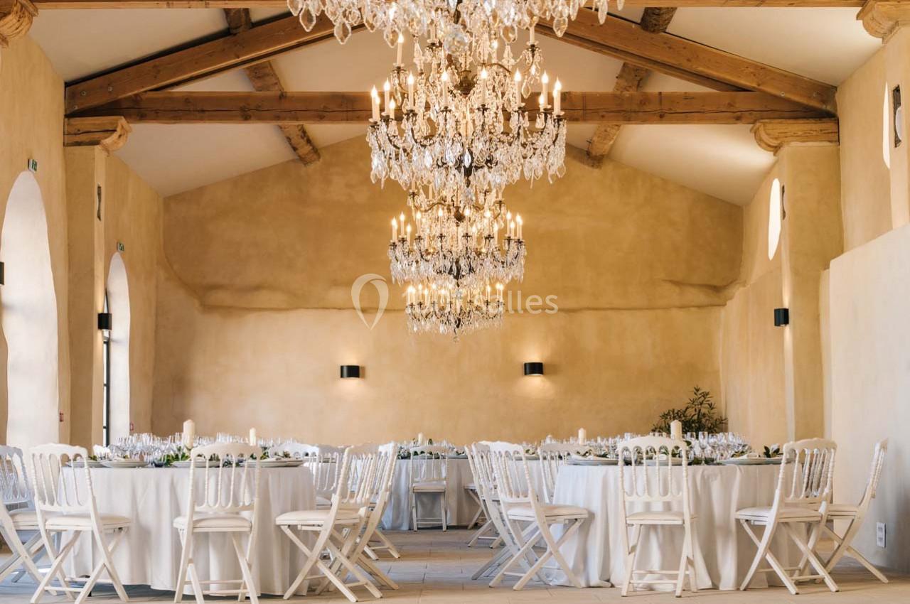 Salle de réception lumineuse avec tables rondes dressées, chaises blanches et lustres en cristal suspendus.