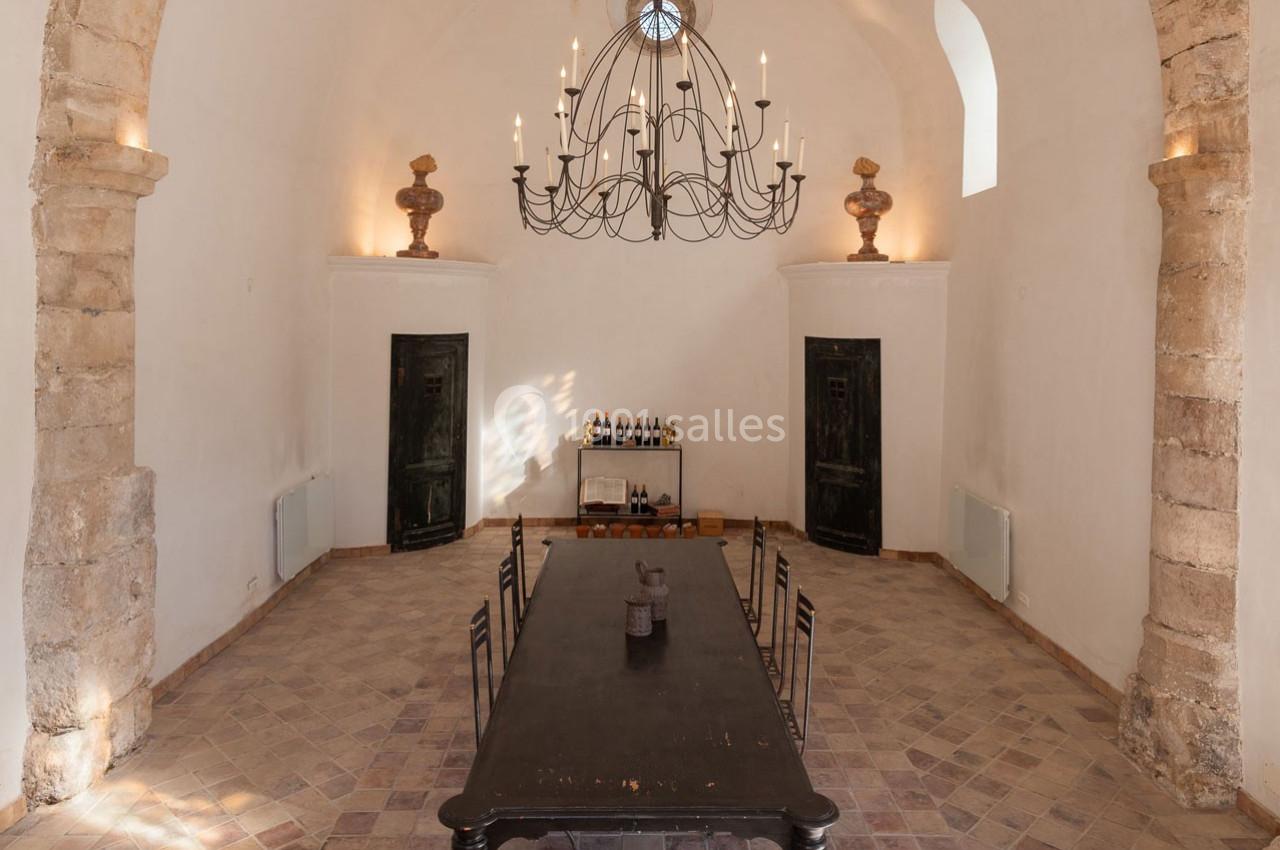 Salle voûtée avec une grande table en bois sombre, un lustre en fer forgé et des murs en pierre claire.