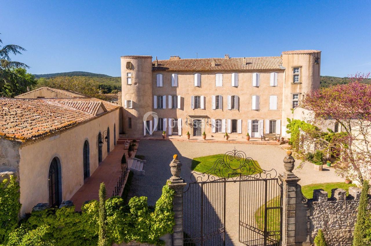 Façade d'un château historique entouré d'une cour pavée, avec jardin et portail en fer forgé, sous un ciel dégagé.