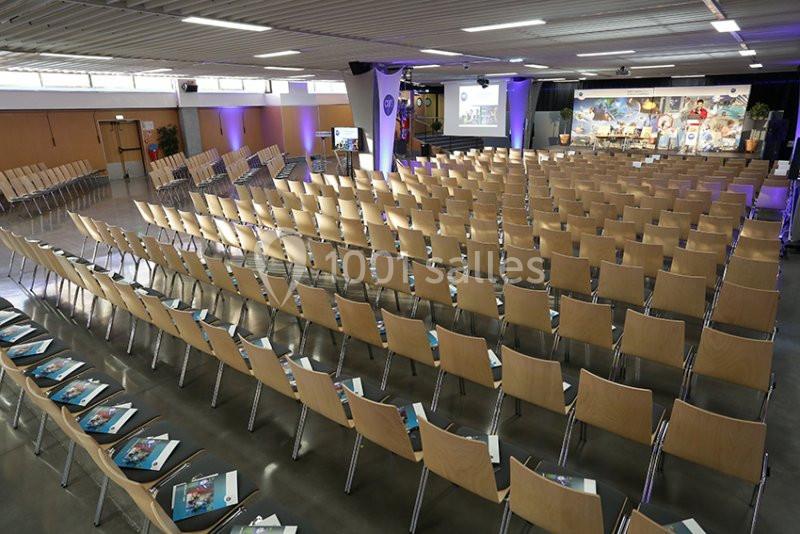 Salle de conférence avec rangées de chaises en bois, brochures posées sur certaines, et une scène équipée en fond.