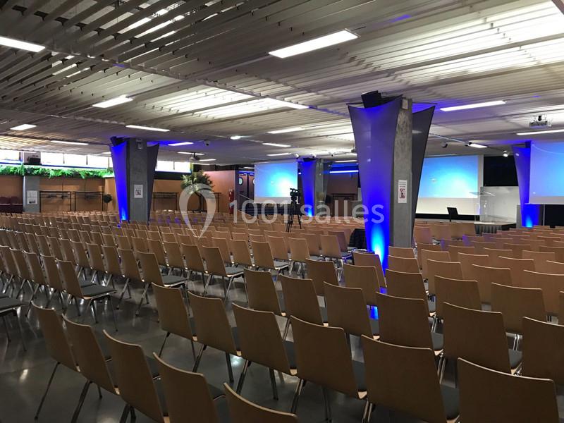 Salle de conférence vide avec rangées de chaises en bois, écrans de projection et éclairage bleu.