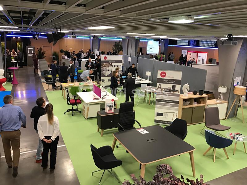 Vue d'un salon professionnel avec stands de mobilier de bureau, visiteurs et espaces d'exposition modernes.