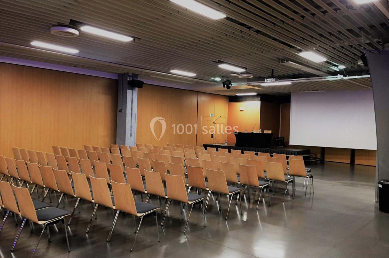Salle de conférence vide avec rangées de chaises en bois, un écran de projection et une table pour les intervenants.