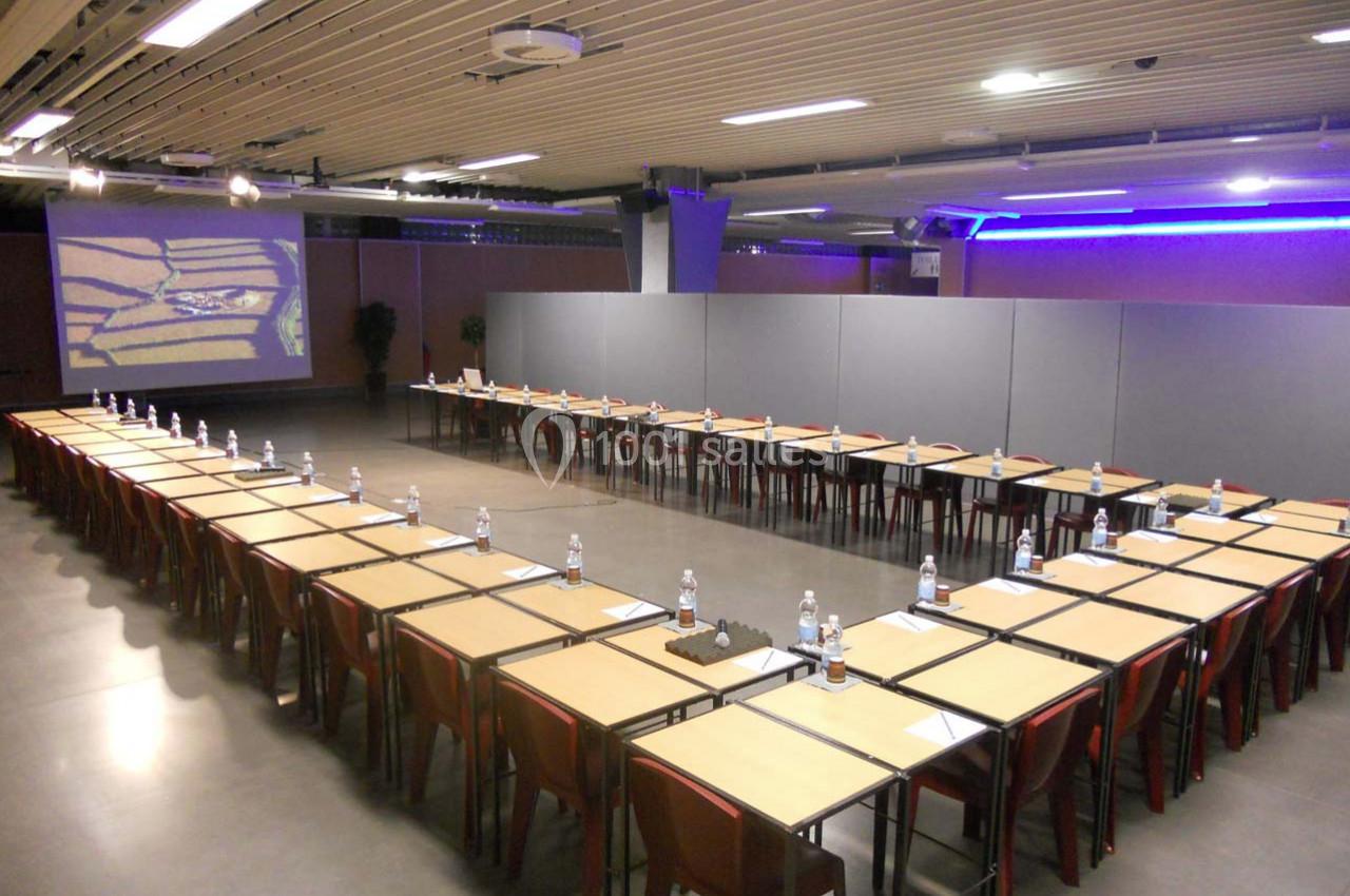 Salle de conférence avec des tables disposées en U, équipées de bouteilles d'eau et de documents, éclairage moderne.