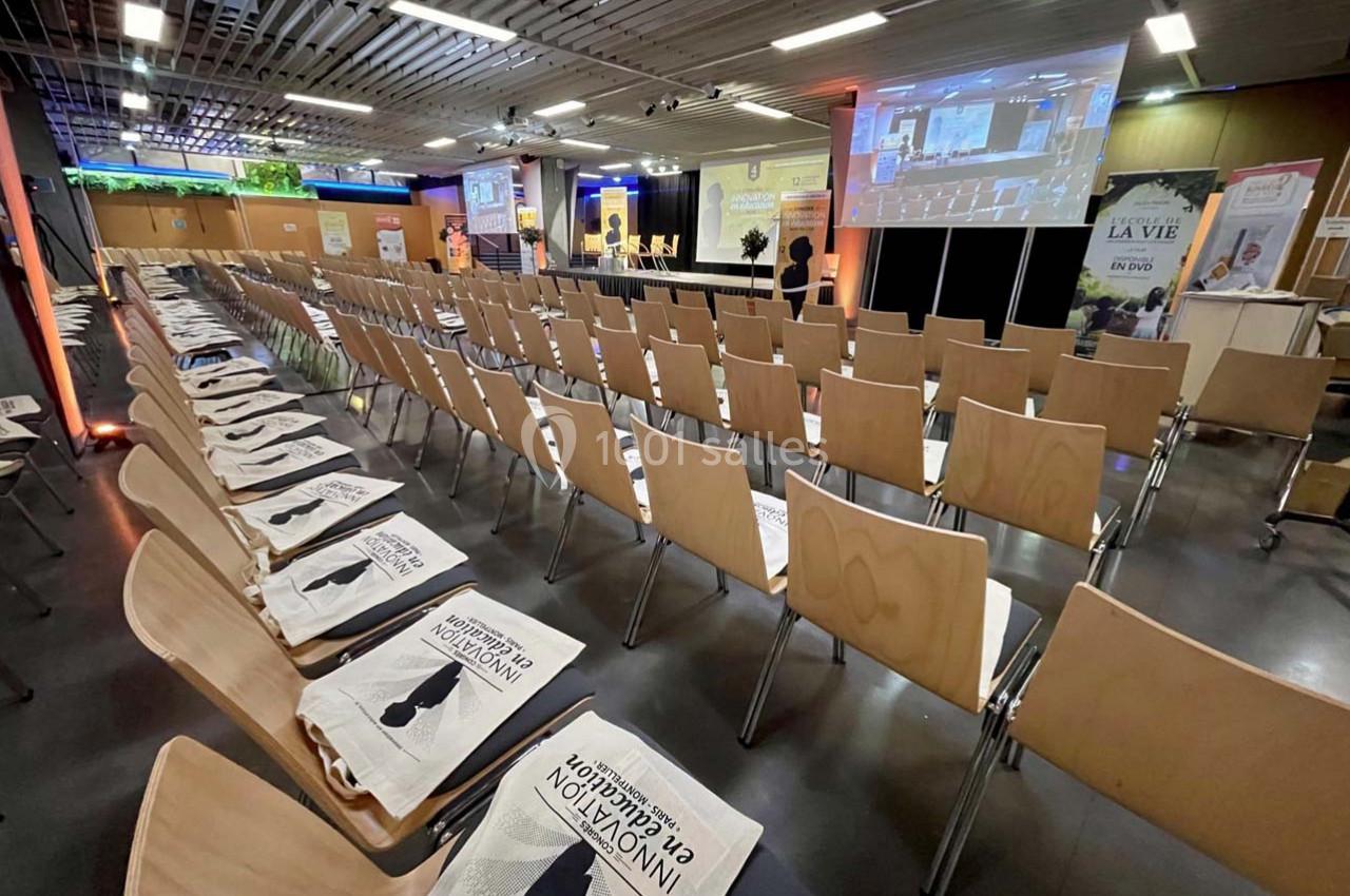 Salle de conférence avec des rangées de chaises en bois, sacs promotionnels posés dessus, et une scène équipée d'écrans.