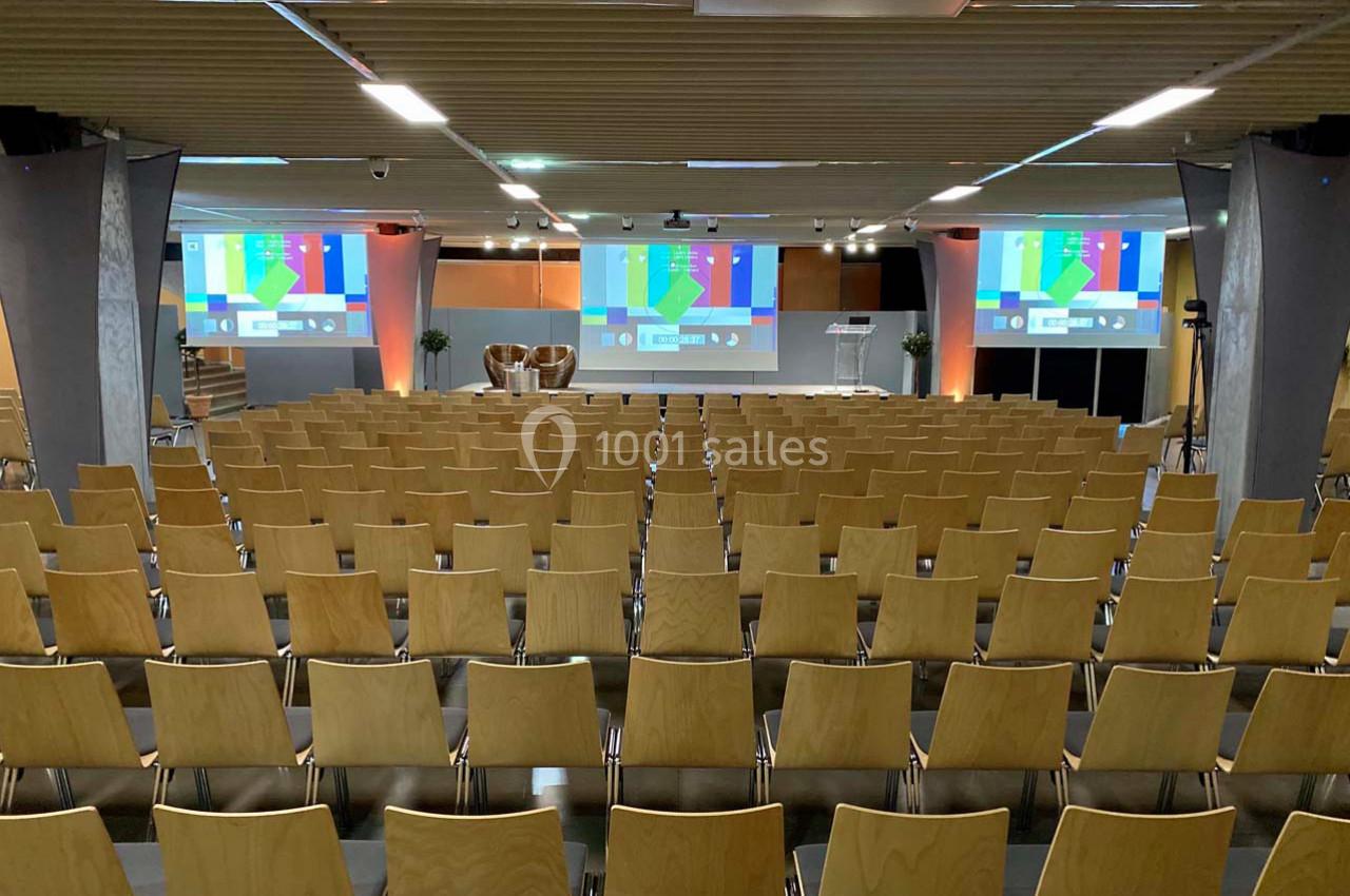 Salle de conférence vide avec des rangées de chaises en bois, un écran de projection et un éclairage tamisé.