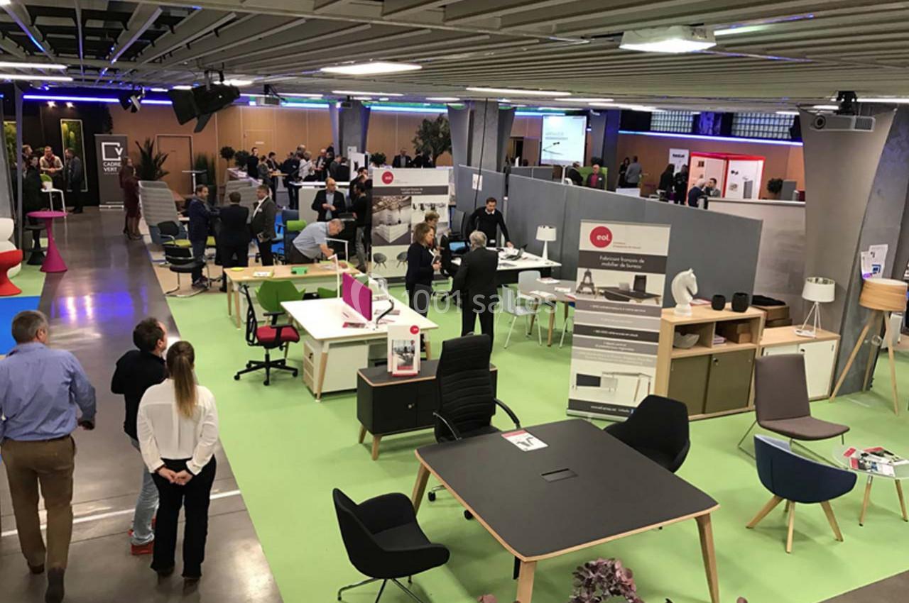 Vue d'un salon professionnel avec des stands présentant du mobilier de bureau et des visiteurs échangeant.