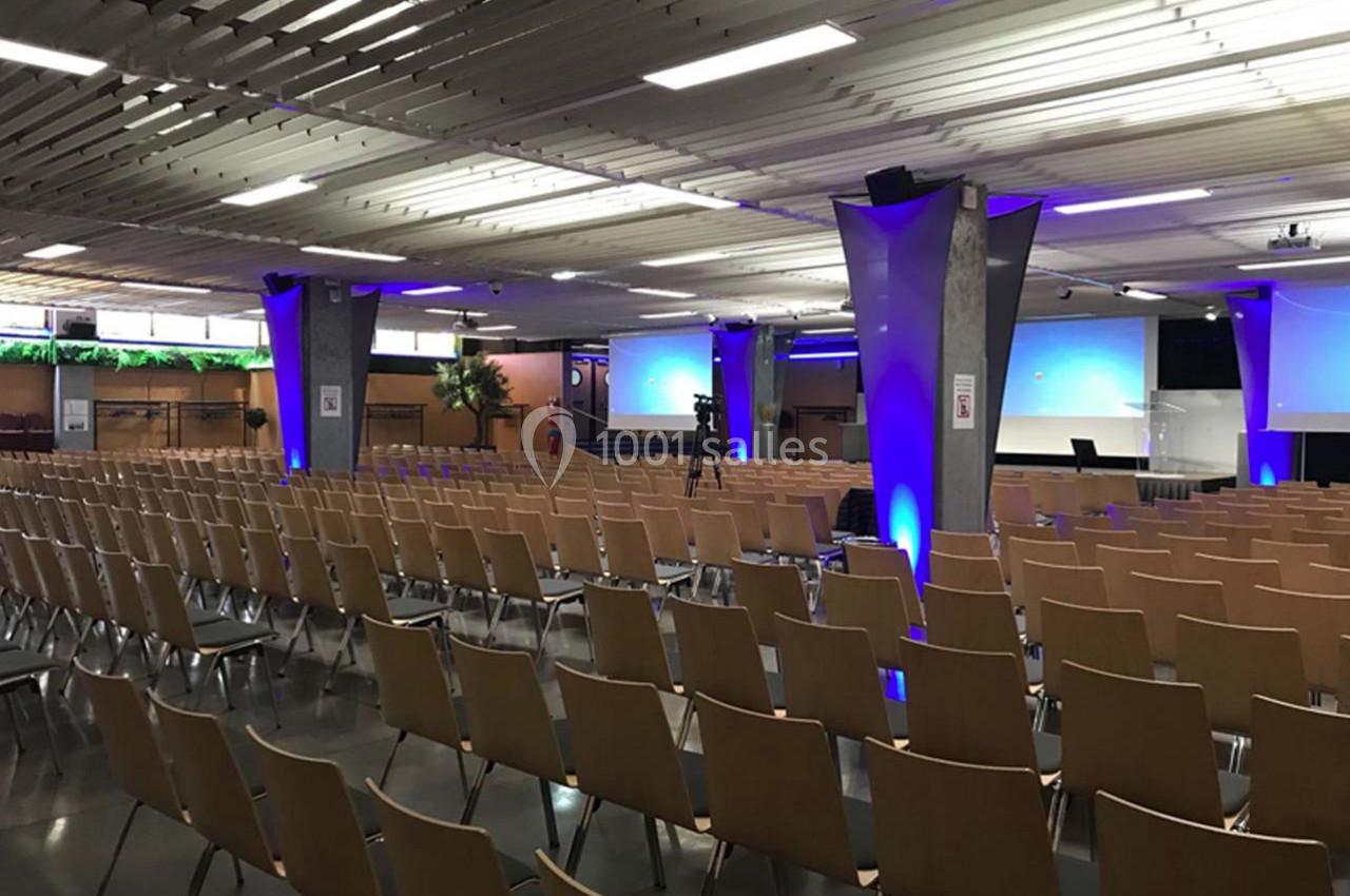 Salle de conférence vide avec rangées de chaises en bois, écrans de projection et éclairage bleu.