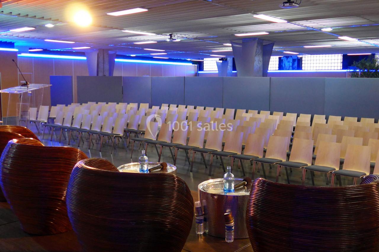 Salle de conférence vide avec rangées de chaises en bois, vue depuis une estrade équipée de tables et bouteilles d'eau.