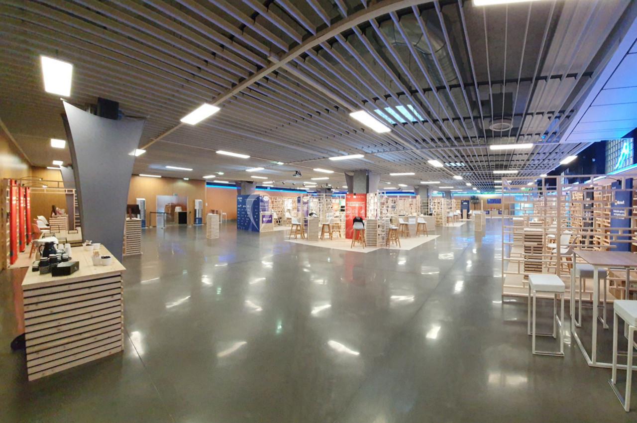 Salle spacieuse avec un sol en béton, des structures en bois, des tables hautes et un éclairage fluorescent.