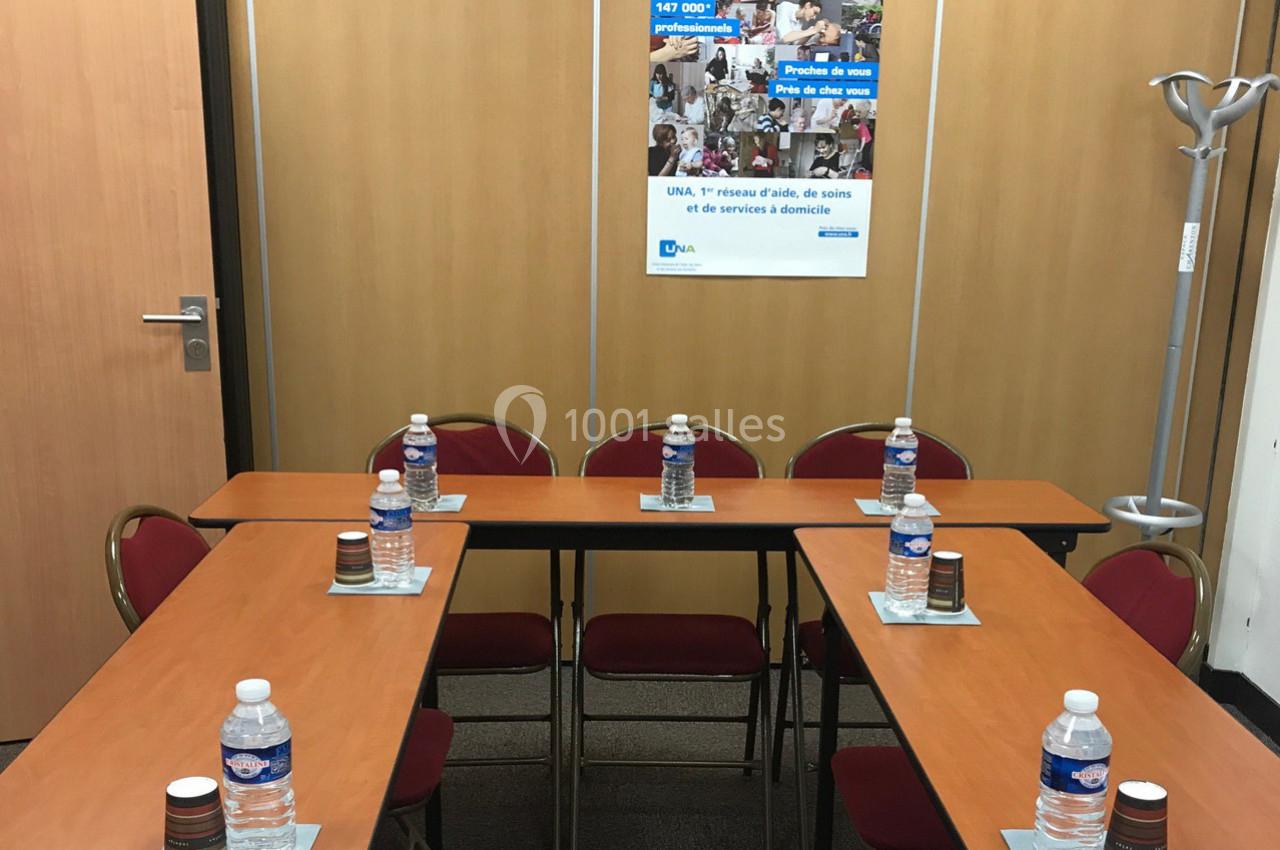 Salle de réunion avec tables disposées en U, bouteilles d'eau et gobelets, panneau d'information en arrière-plan.