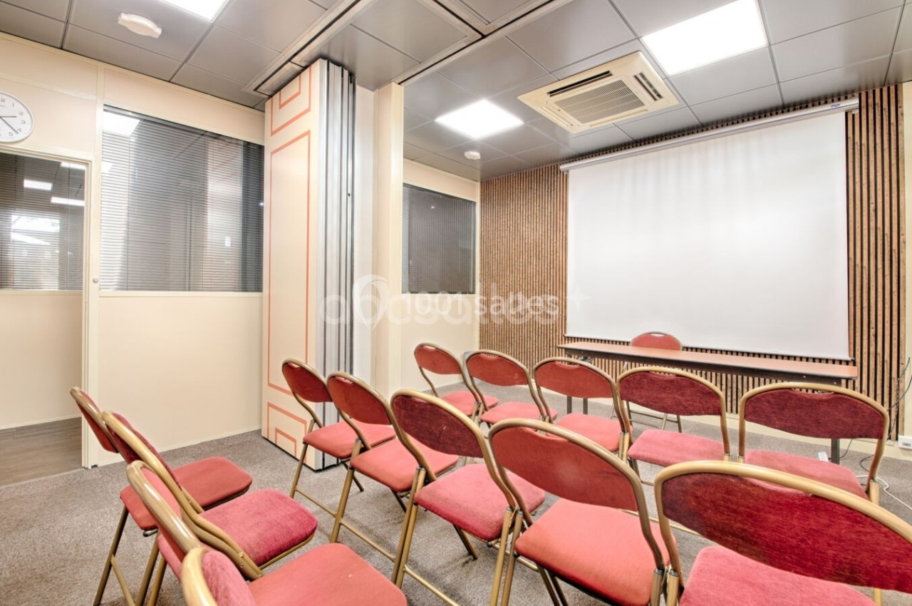 Salle de réunion avec chaises rouges alignées face à un écran de projection dans un espace lumineux et moderne.