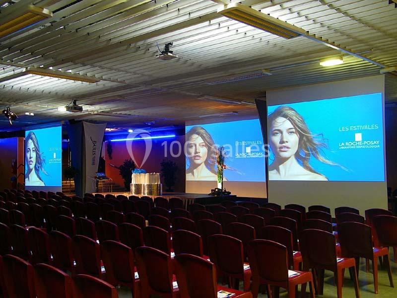 Salle de conférence avec rangées de chaises rouges et écrans affichant une présentation intitulée ’Les Estivales’.