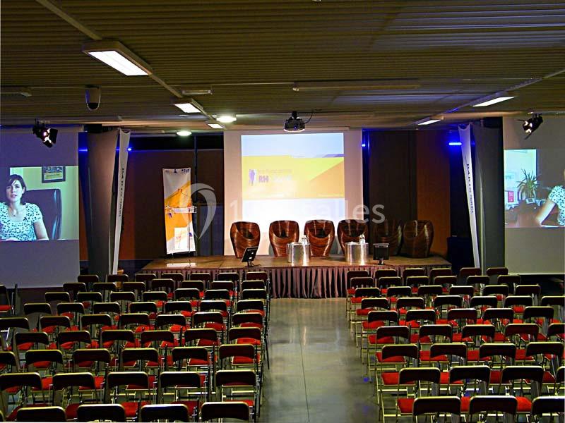 Salle de conférence vide avec des rangées de chaises, une scène équipée de fauteuils et des écrans affichant des…