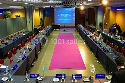 Salle de réunion avec tables disposées en U, bouteilles d'eau et gobelets, panneau d'information en arrière-plan.