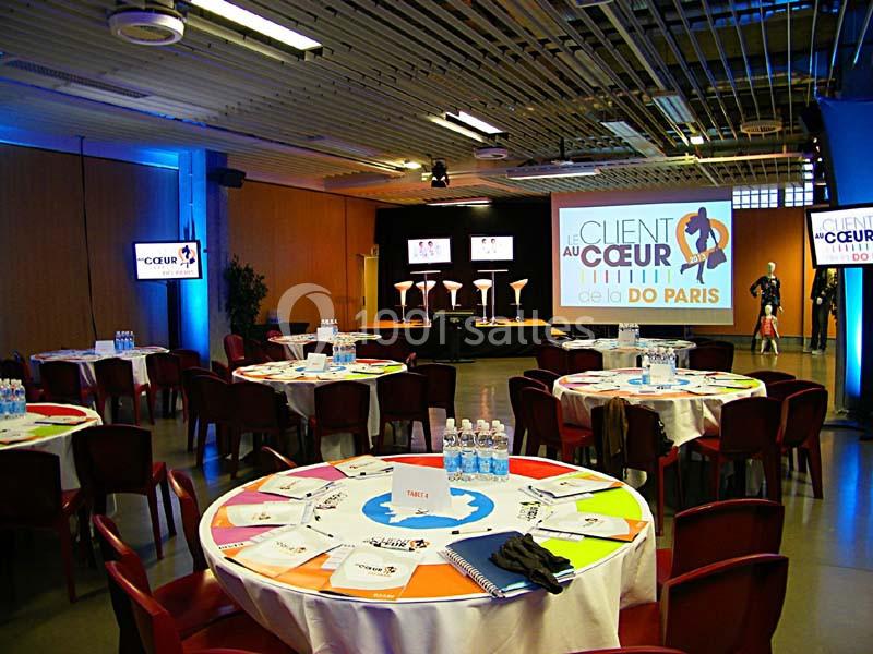 Salle de conférence avec tables rondes dressées, documents et bouteilles d'eau, scène équipée pour une présentation.