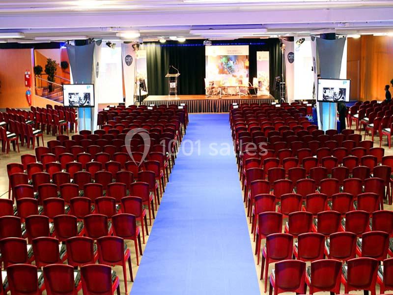 Salle de conférence avec des rangées de chaises rouges, une allée centrale bleue et une scène équipée d'un pupitre.