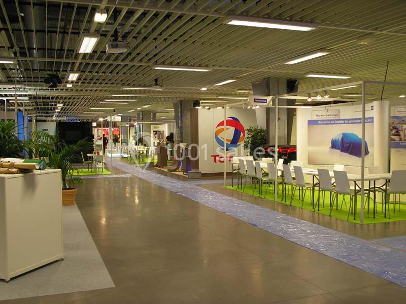 Salle d'exposition avec stands, tables blanches, plantes et éclairage fluorescent sous un plafond métallique.