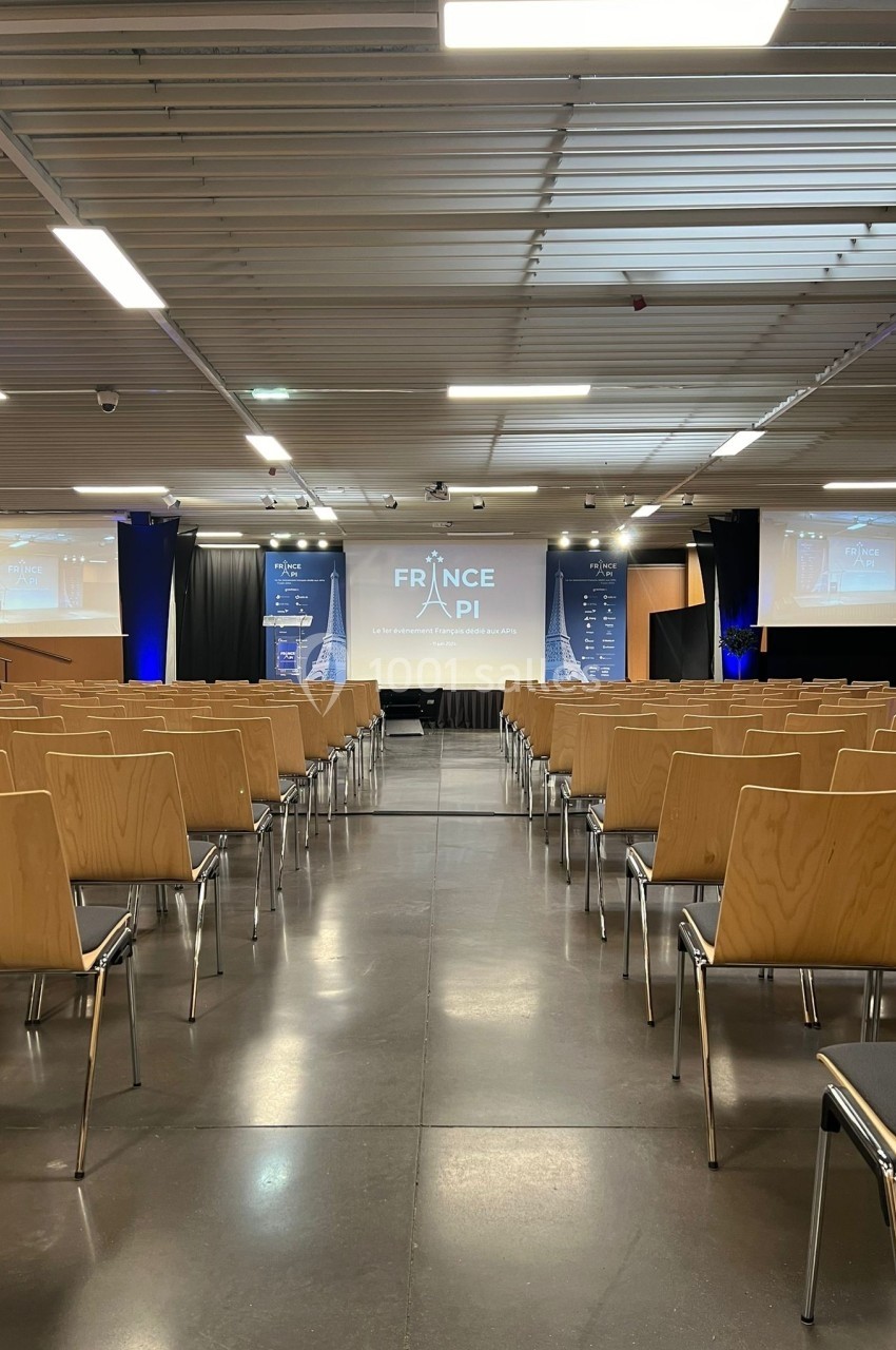 Salle de conférence vide avec des rangées de chaises en bois, un écran principal et deux écrans latéraux affichant ’France…