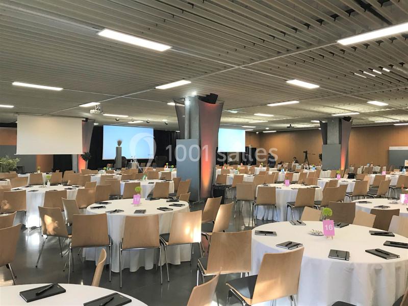 Salle de conférence avec des tables rondes dressées, des chaises en bois et des écrans de projection en arrière-plan.