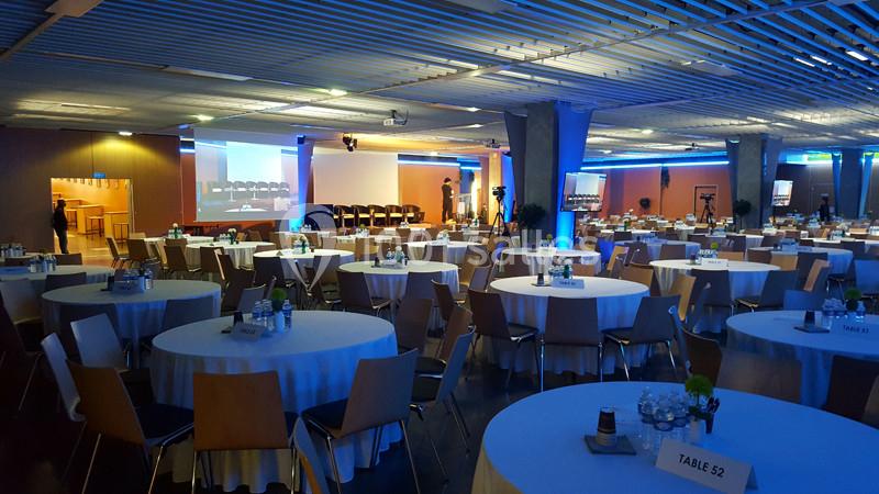 Salle de conférence aménagée avec des tables rondes, des chaises et un écran de projection allumé.