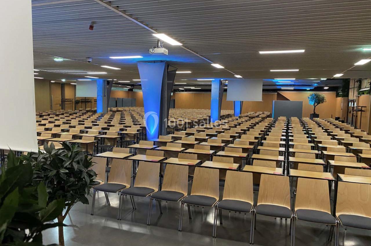 Salle de conférence vide avec rangées de chaises en bois et écrans de projection, éclairage bleu sur les colonnes.