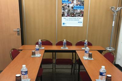Salle de réunion avec tables disposées en U, bouteilles d'eau et gobelets, panneau d'information en arrière-plan.
