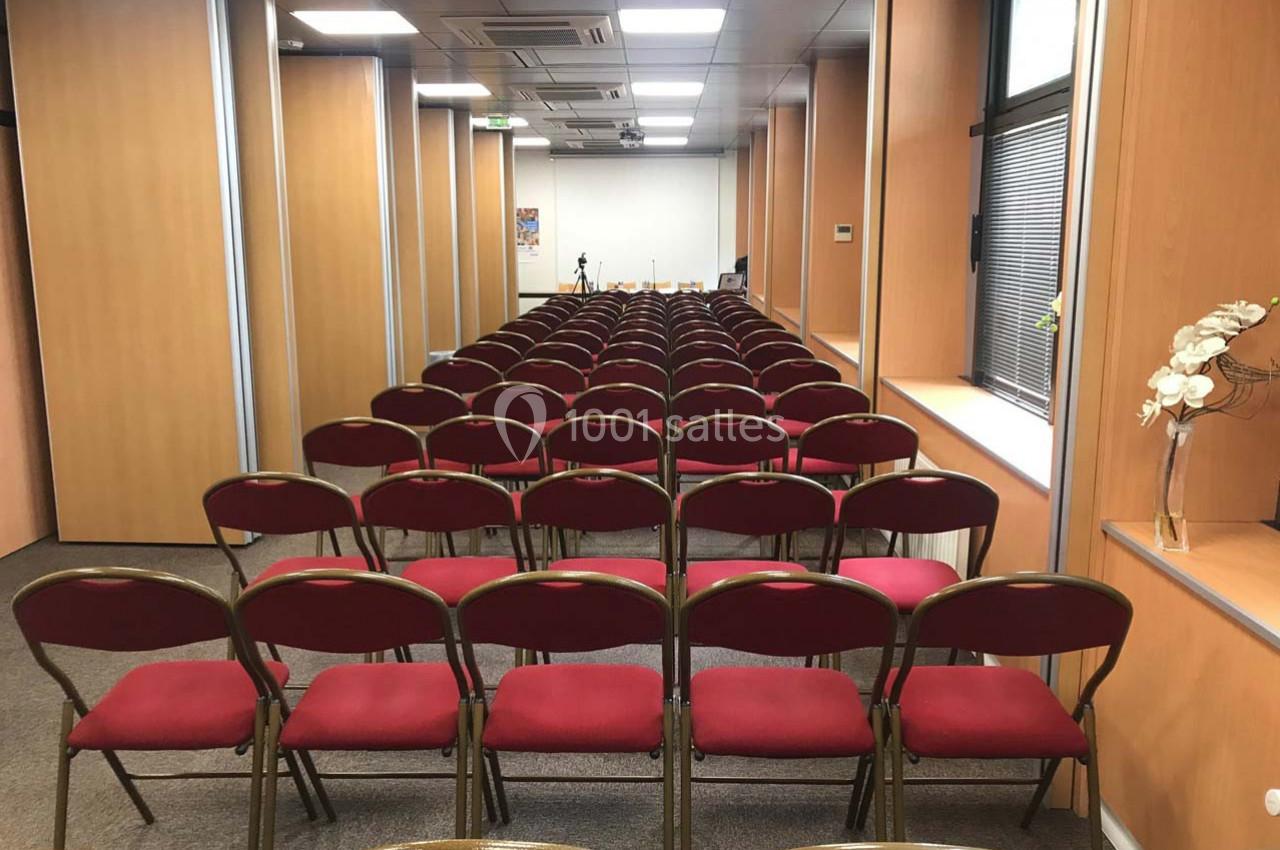 Salle de conférence avec rangées de chaises rouges alignées face à un écran blanc.