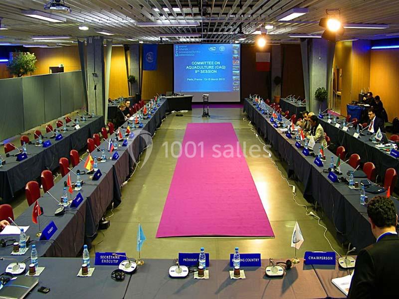 Salle de conférence avec une longue table centrale entourée de chaises, écrans et drapeaux représentant différents pays.