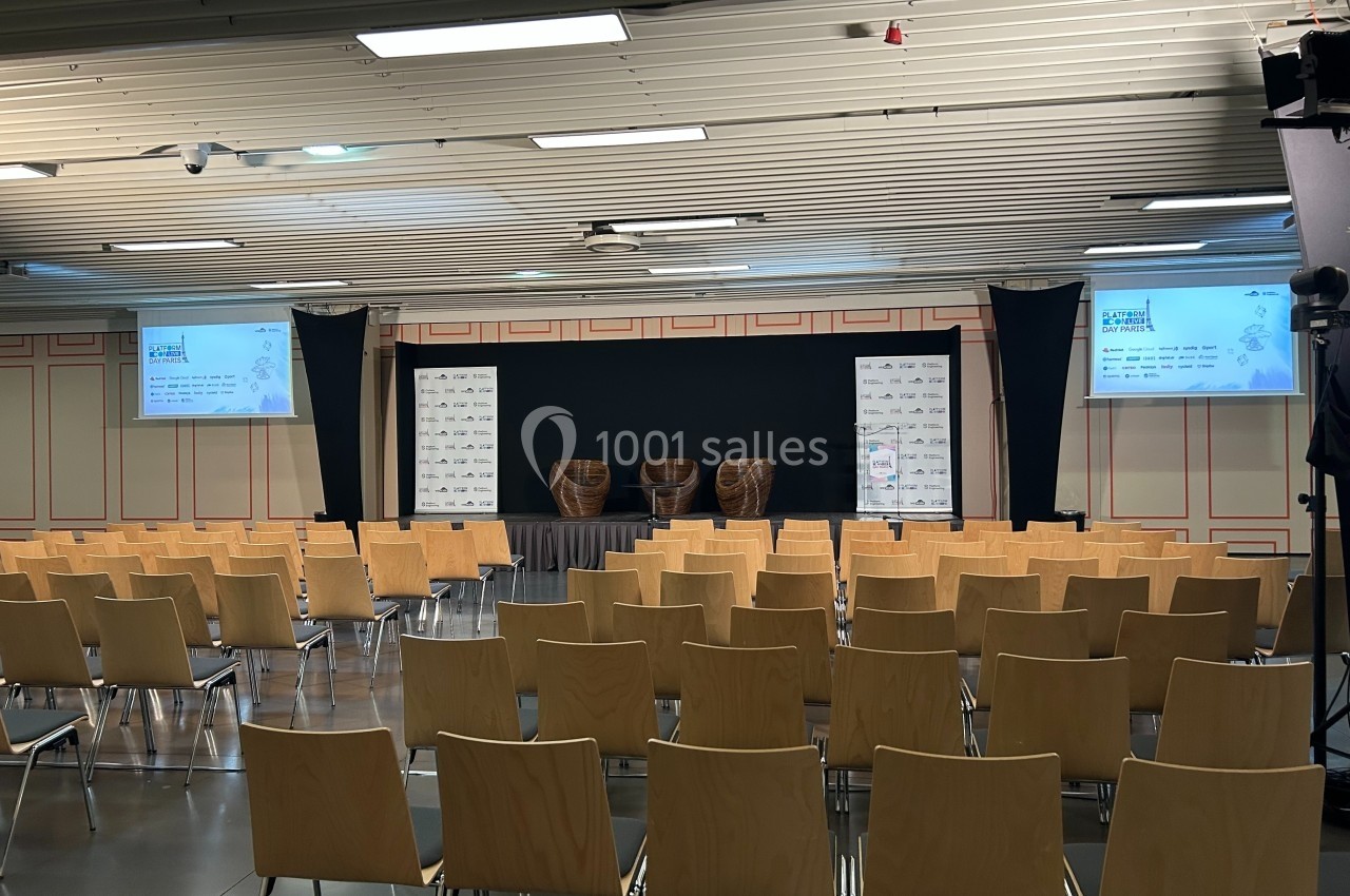 Salle de conférence vide avec des rangées de chaises en bois, une scène équipée de deux fauteuils et des écrans affichant…