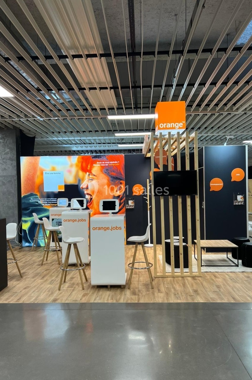 Stand d'exposition avec des écrans, des affiches et des meubles aux couleurs de l'entreprise Orange.