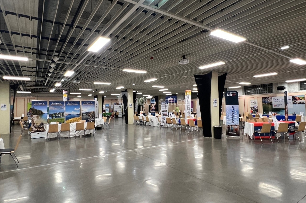 Salle d'exposition avec des stands alignés sous un plafond industriel, éclairée par des lumières artificielles.
