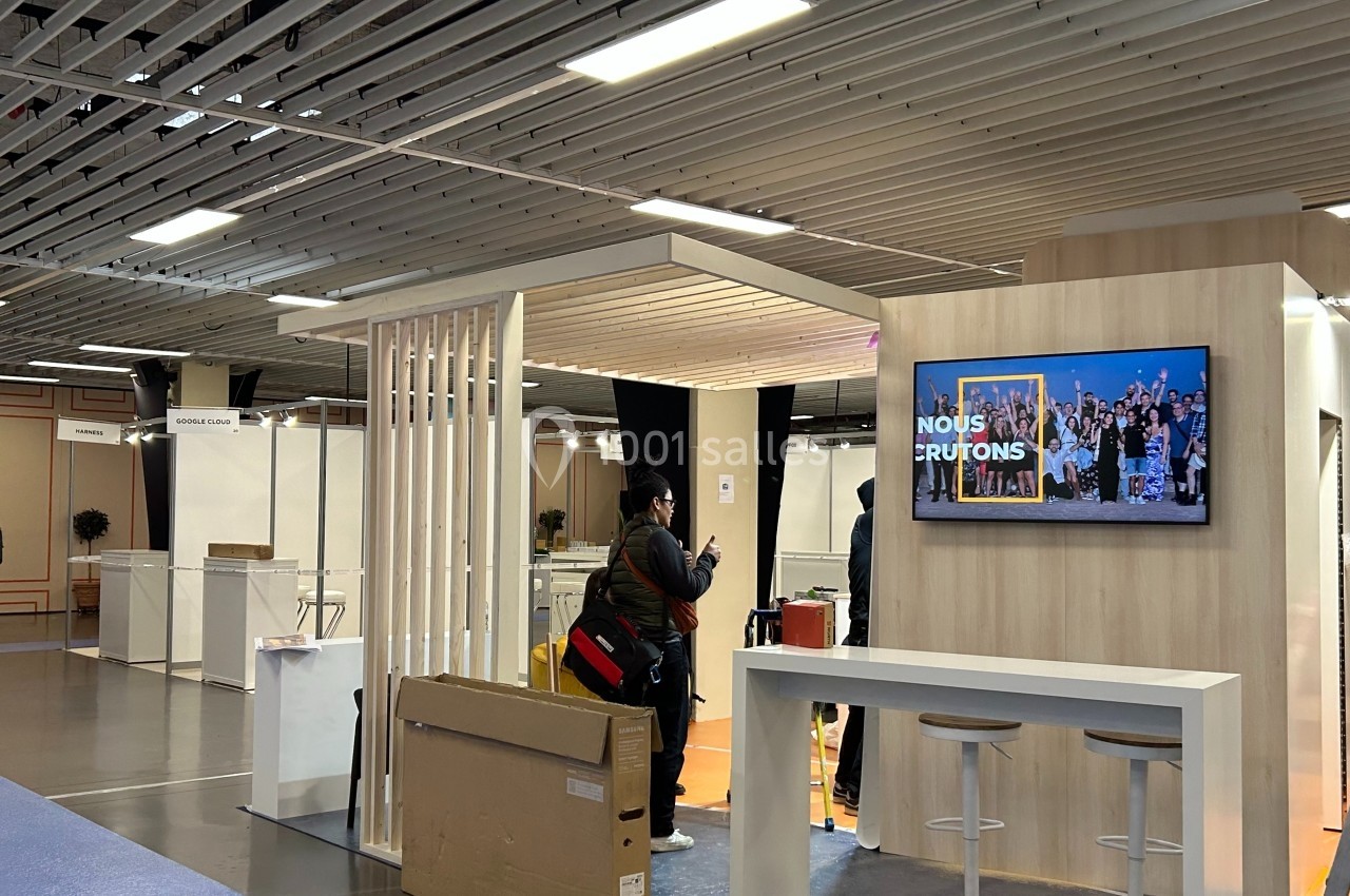 Stand d'exposition avec un écran affichant ’Nous recrutons’, des tabourets hauts et une personne prenant une photo.