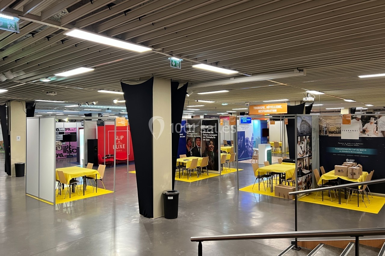 Salle d'exposition avec plusieurs stands colorés, tables jaunes et éclairage fluorescent.