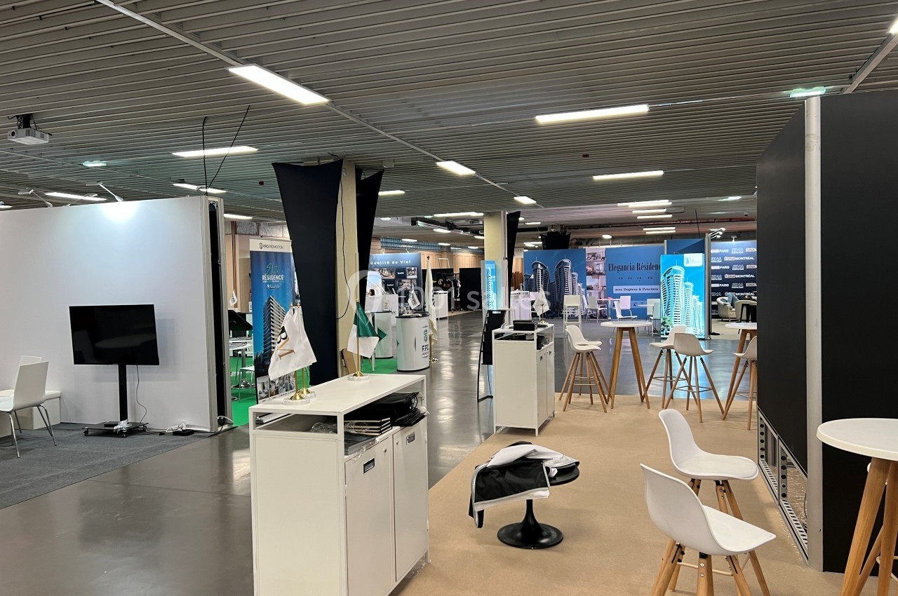Espace d'exposition intérieur avec stands, maquettes architecturales, chaises blanches et éclairage fluorescent.