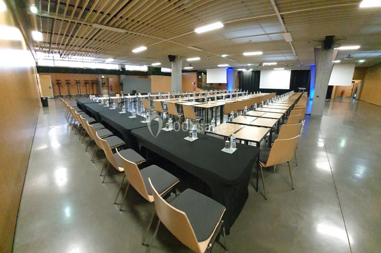 Salle de conférence avec des rangées de tables et chaises alignées, équipée de bouteilles d'eau et de verres.