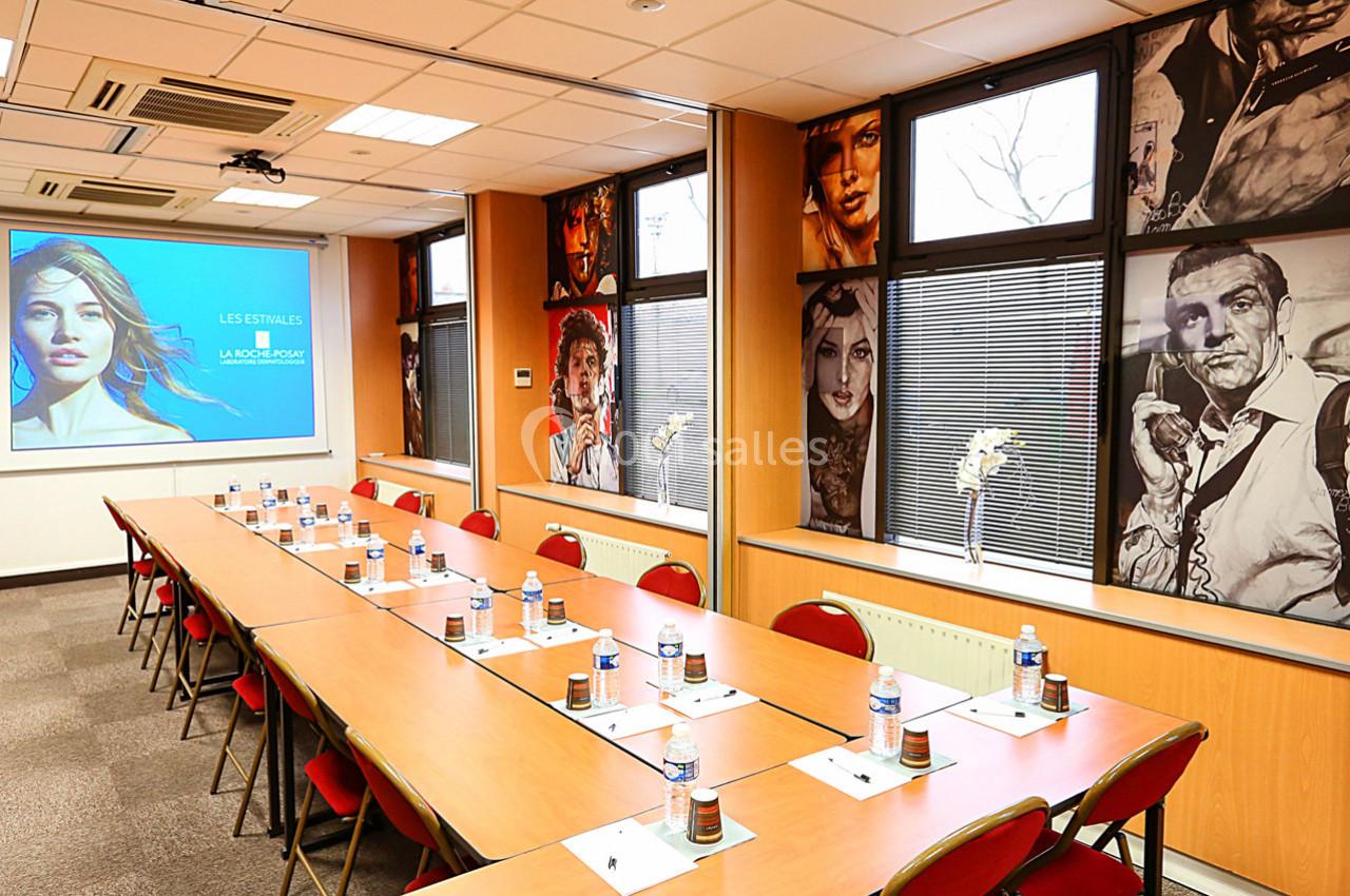 Salle de réunion équipée avec tables en U, bouteilles d'eau, bloc-notes, écran de projection et décorations murales.