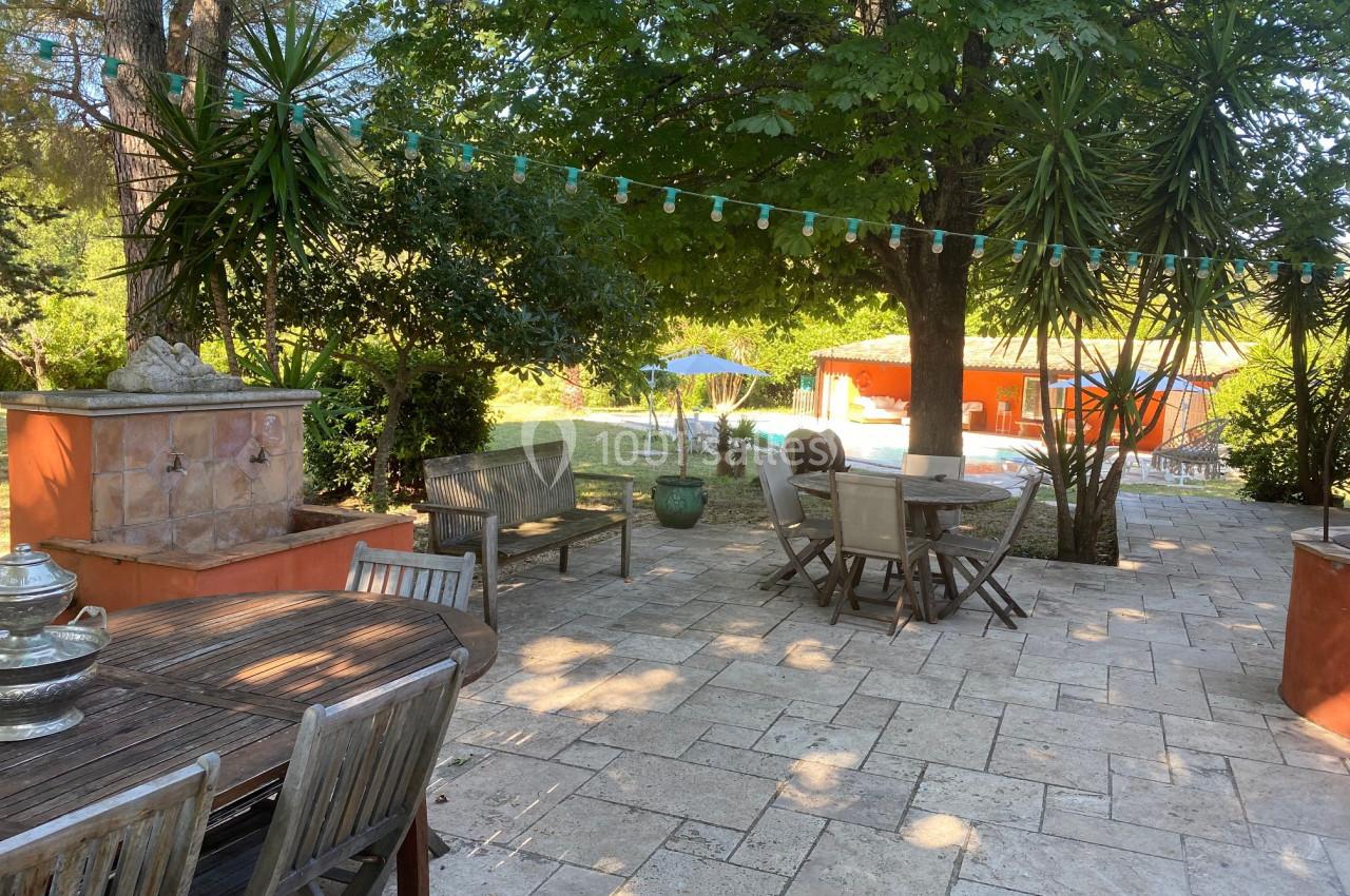 Terrasse en pierre ombragée avec mobilier de jardin, guirlande lumineuse et vue sur une piscine entourée de verdure.