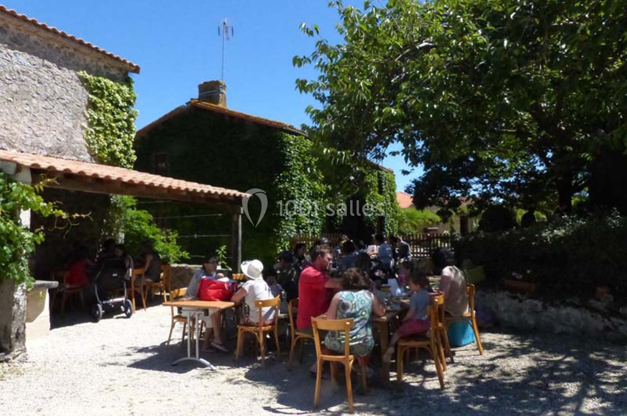 Des personnes assises à des tables en plein air devant des bâtiments en pierre couverts de végétation, par une journée…