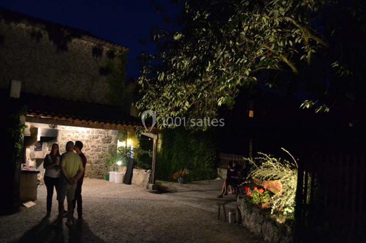 Cour extérieure éclairée de nuit avec un arbre, des plantes et trois personnes discutant près d'une maison en pierre.