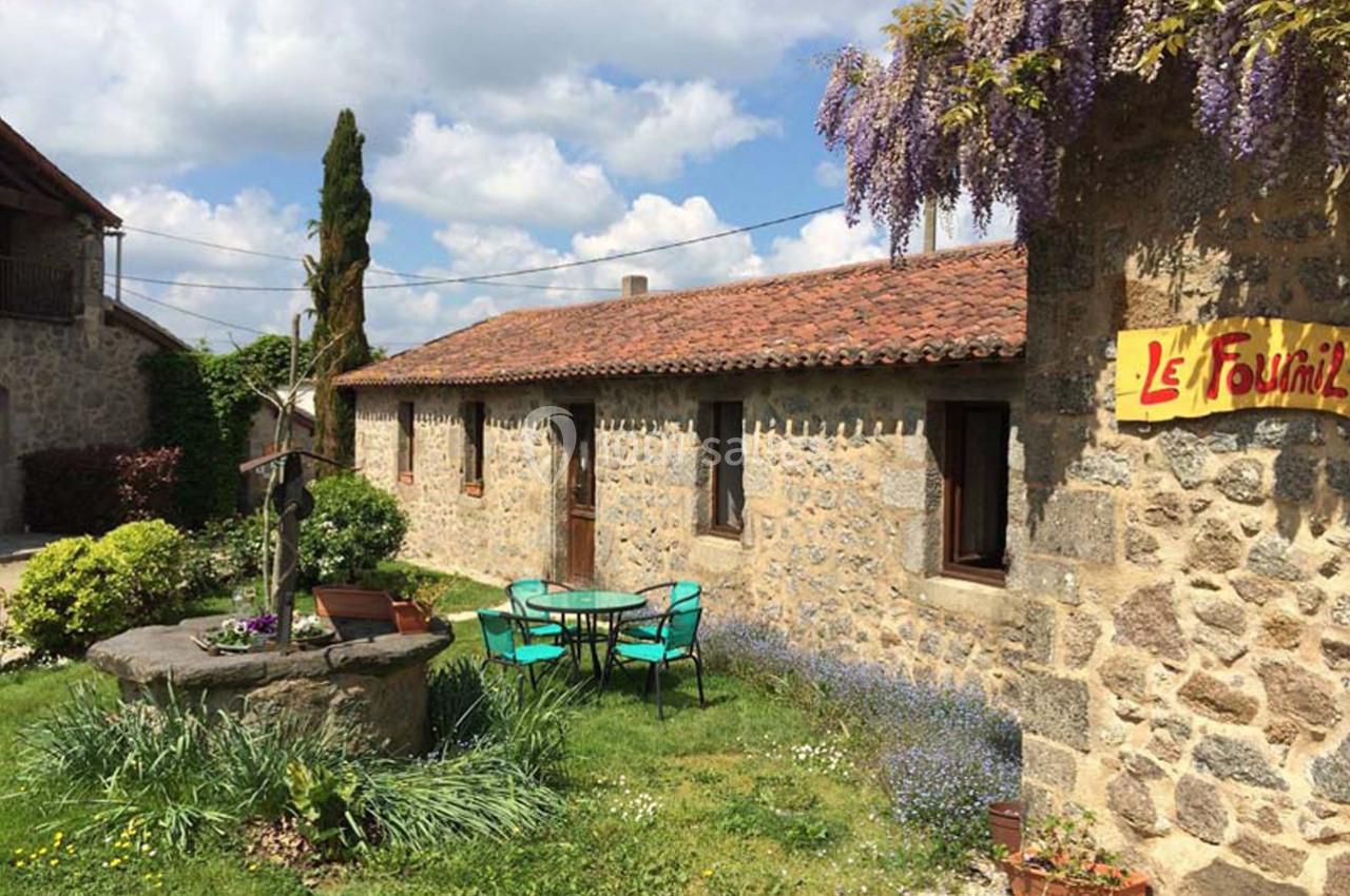 Cour extérieure avec un puits, des chaises et une table, devant une maison en pierre sous un ciel partiellement nuageux.