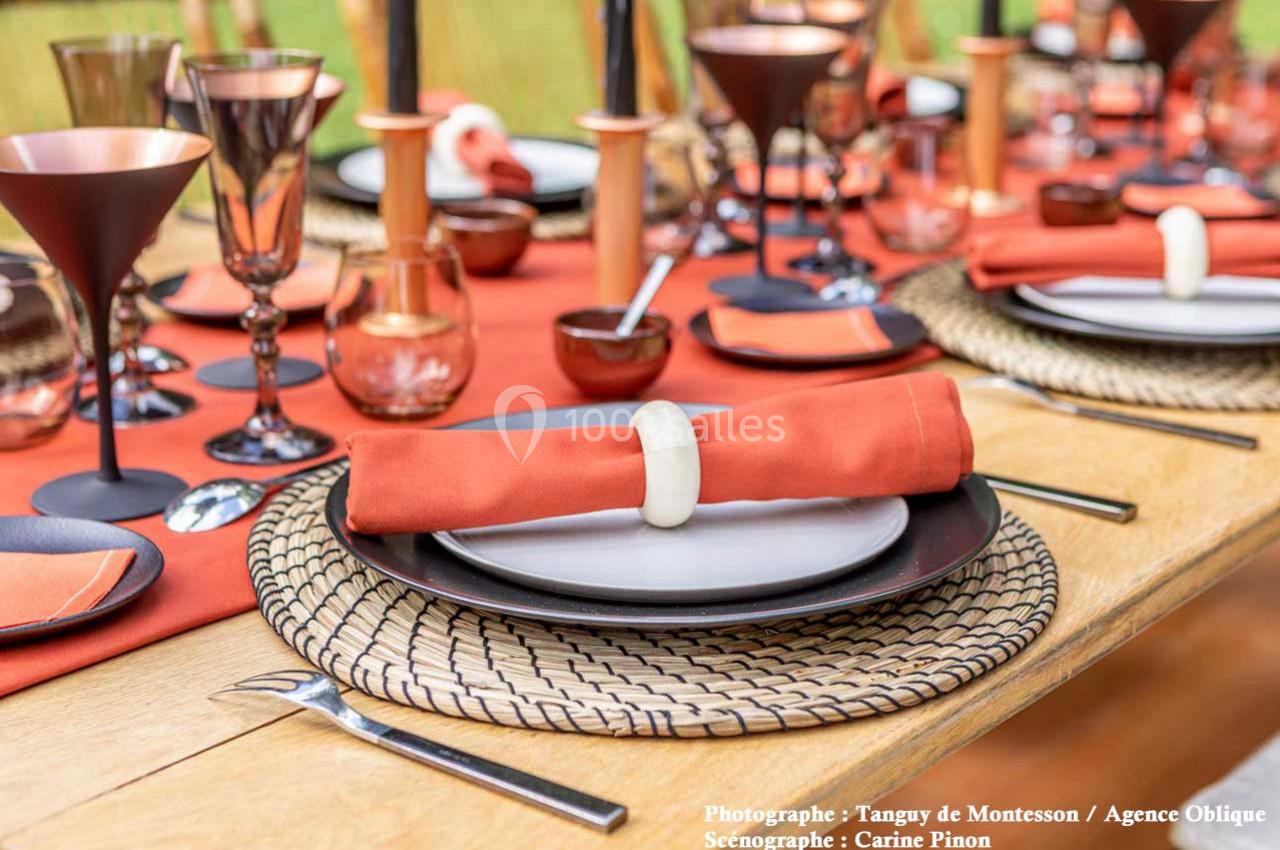 Table dressée avec des assiettes noires, serviettes orange nouées, verres en cuivre et décoration élégante sur nappe rouge.