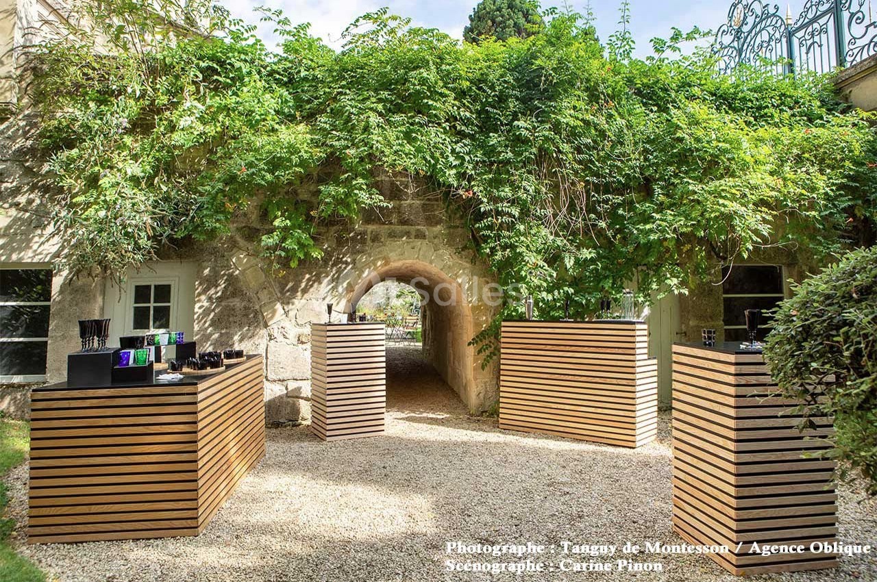Allée extérieure avec arches en pierre, entourée de végétation, et présentoirs en bois alignés sur du gravier.