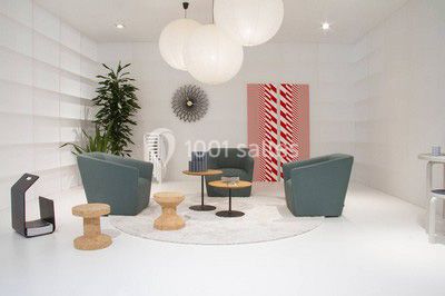 Espace salon moderne avec fauteuils verts, tables basses en bois, tapis rond clair, plantes et décorations murales.