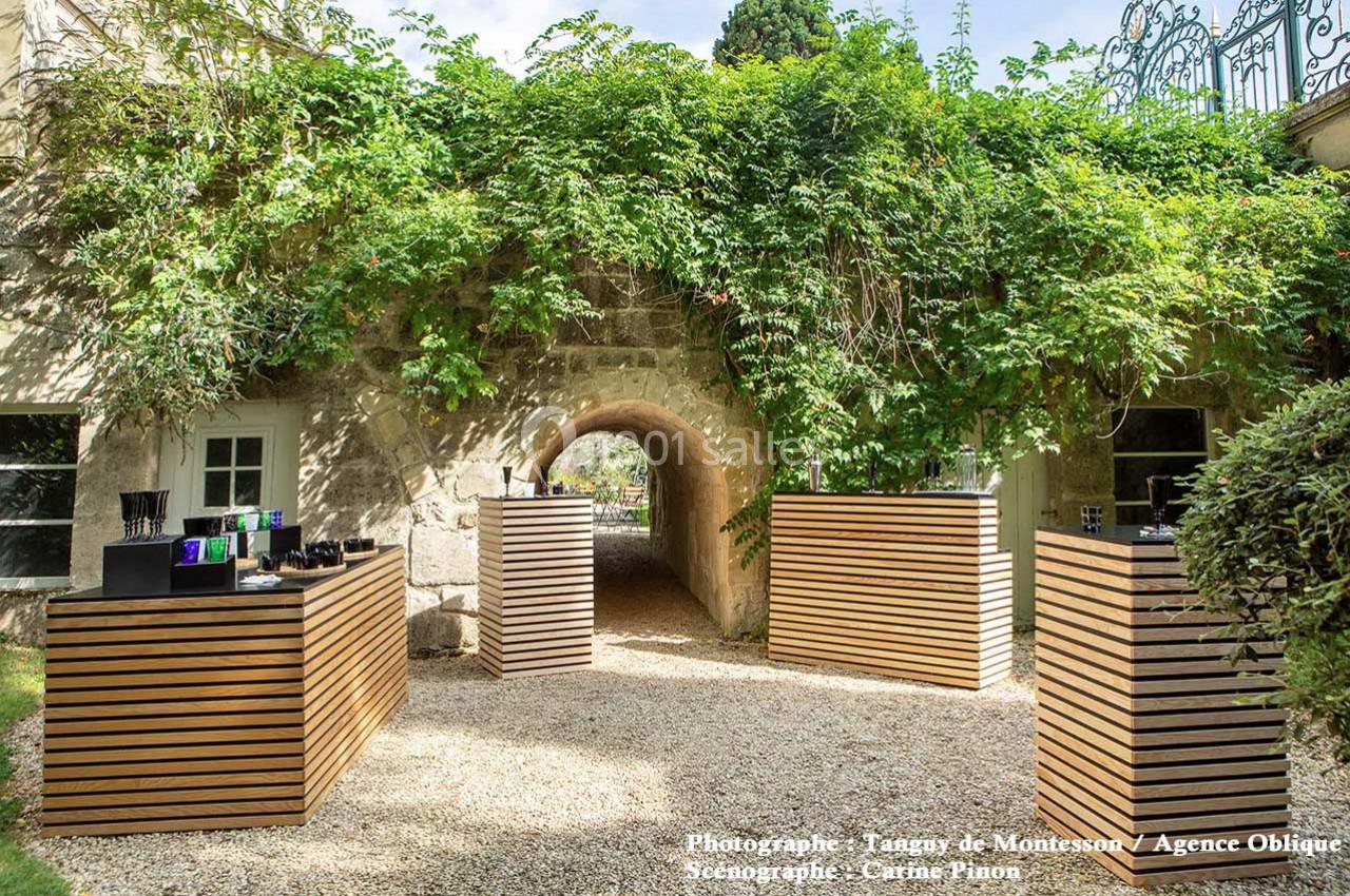 Passage voûté en pierre entouré de végétation, avec des présentoirs en bois alignés sur un chemin gravillonné.